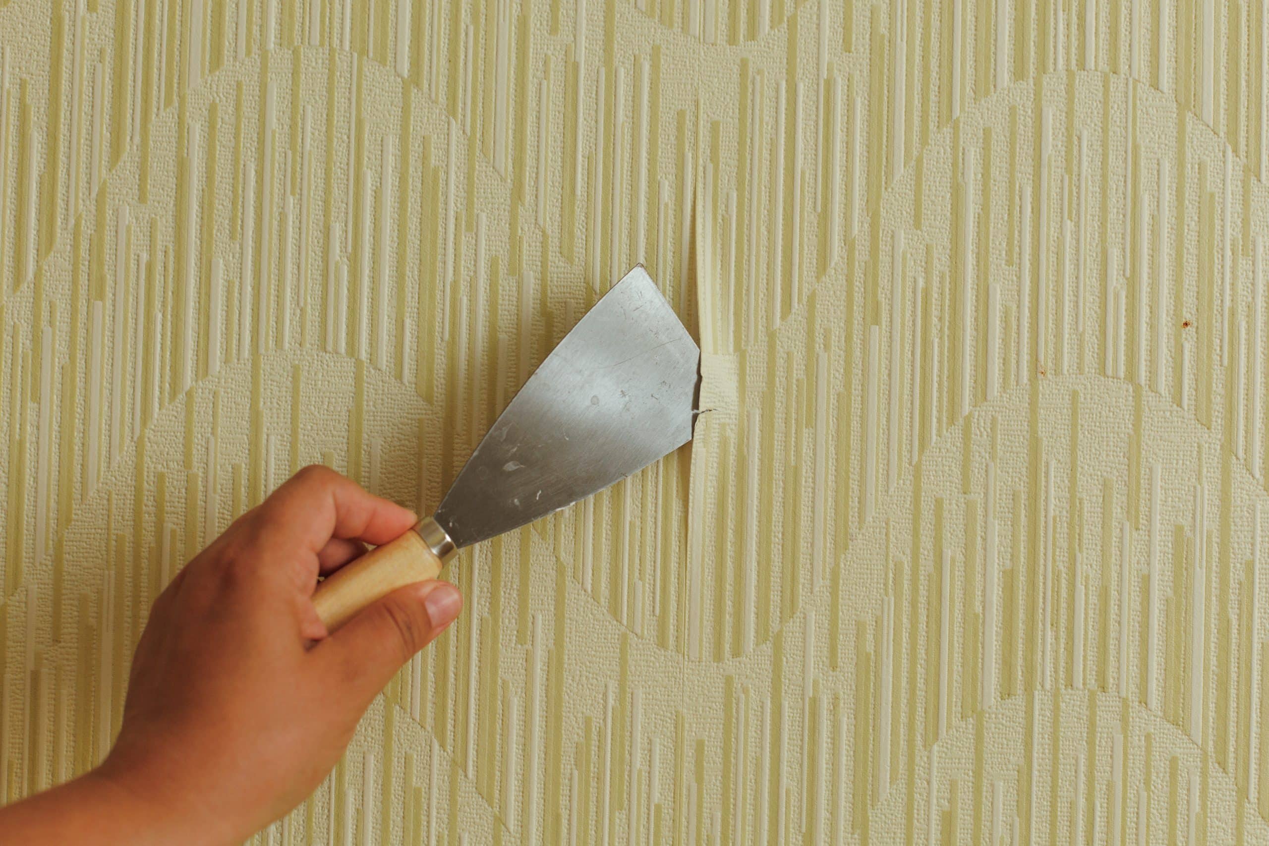 Gloved hands wet-scraping old wallpaper off a wall with a putty knife before the wall is sealed and repainted