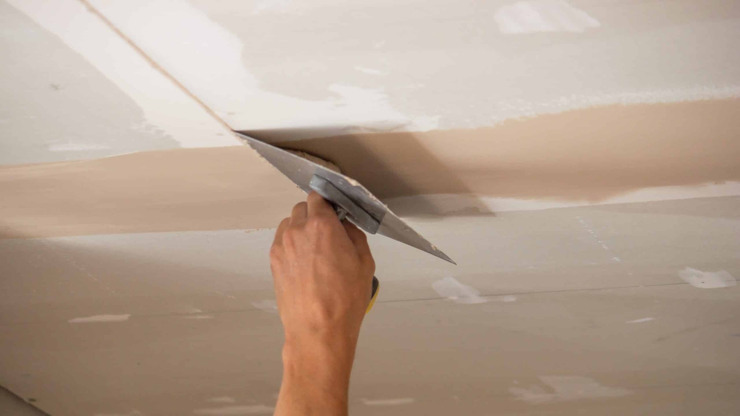 Close-up of joint compound being applied overhead to a plasterboard ceiling seam, showing why the stroke works against gravity on ceiling drywall repair