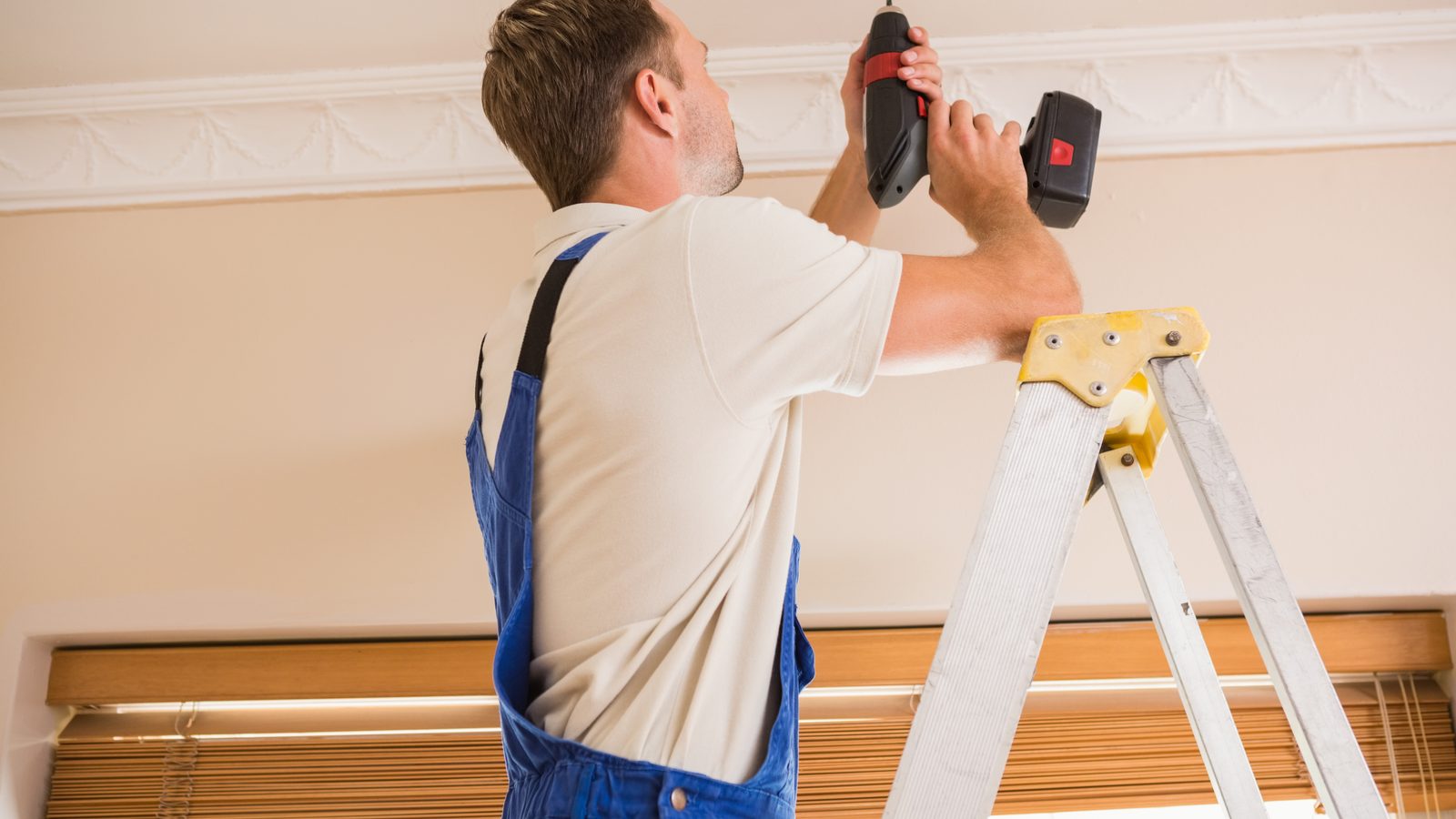 Handyman on a stepladder driving a cordless drill up into a finished ceiling to re-fasten a sagging panel to the joist