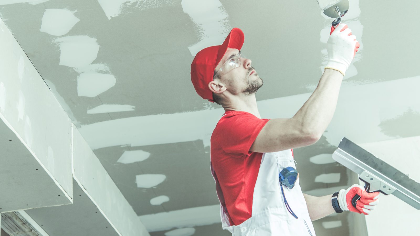 Worker reaching overhead with a wide taping knife to finish the taped seams on an unpainted drywall ceiling.