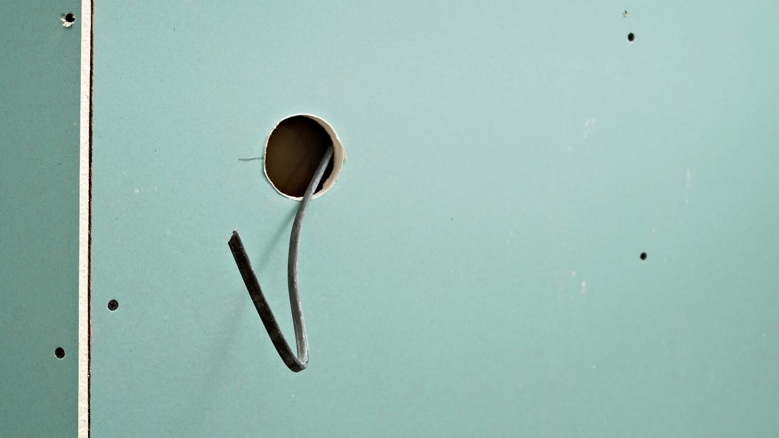 Electrical cable pulled through a round hole cut in a sheet of green drywall, with screw holes nearby