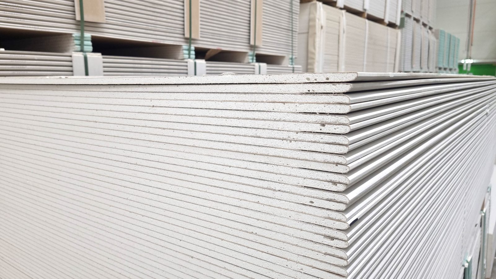 Stack of gypsum drywall sheets seen edge-on in a supply warehouse, showing the board thickness and tapered edges