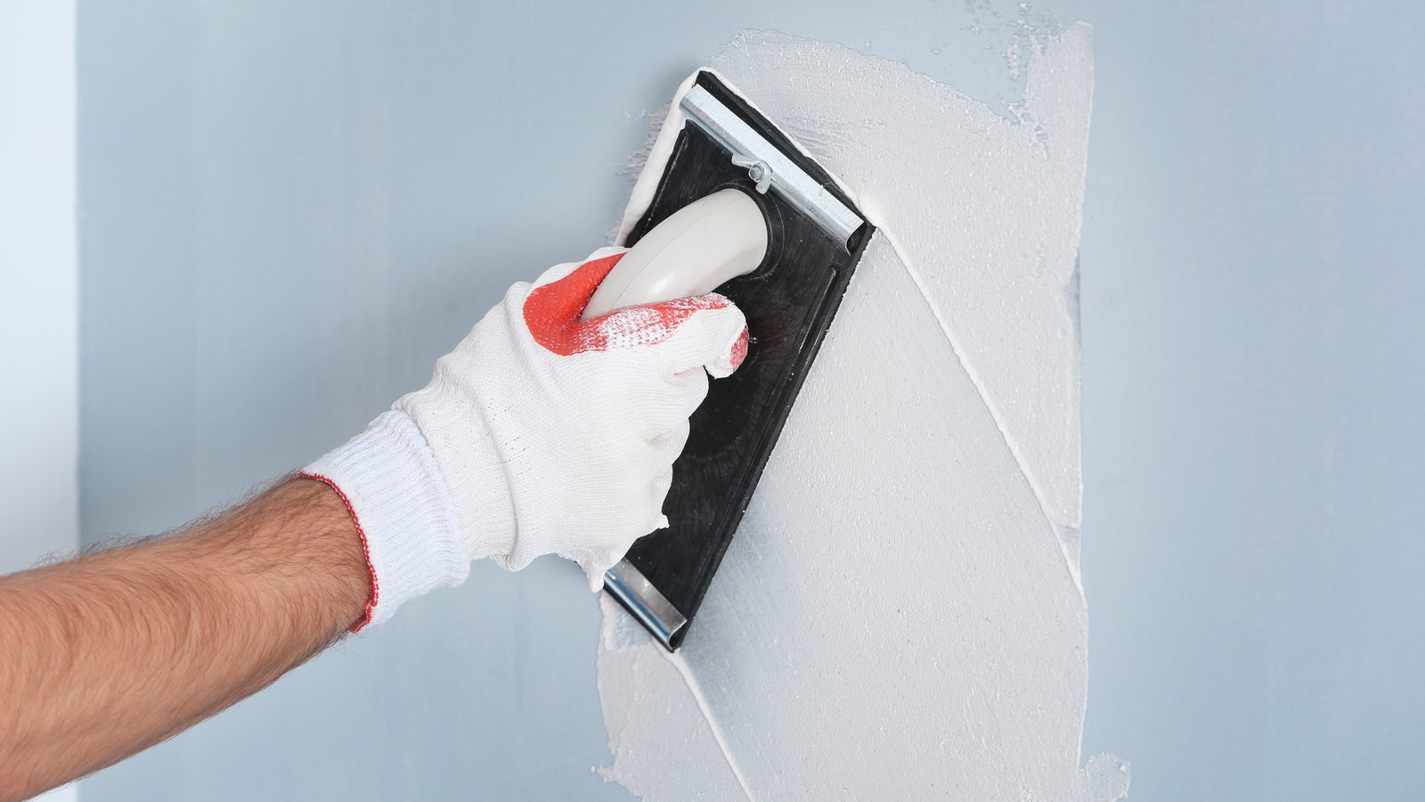 Gloved hand spreading a thin skim coat of joint compound across a drywall surface with a wide finishing knife