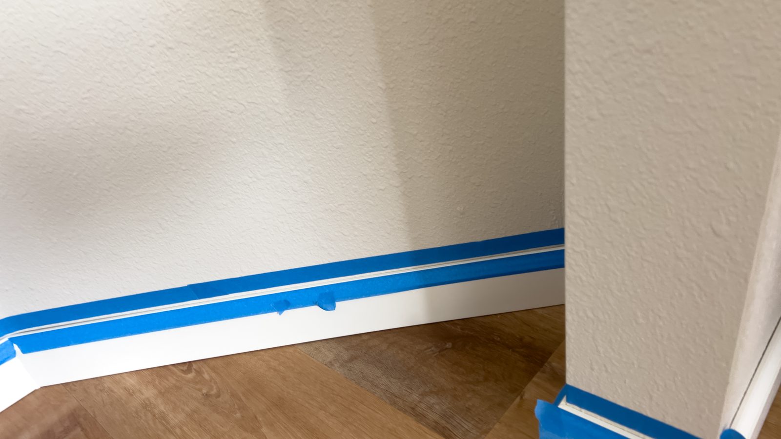 Blue painter's tape run along a white baseboard beneath a lightly textured wall before painting