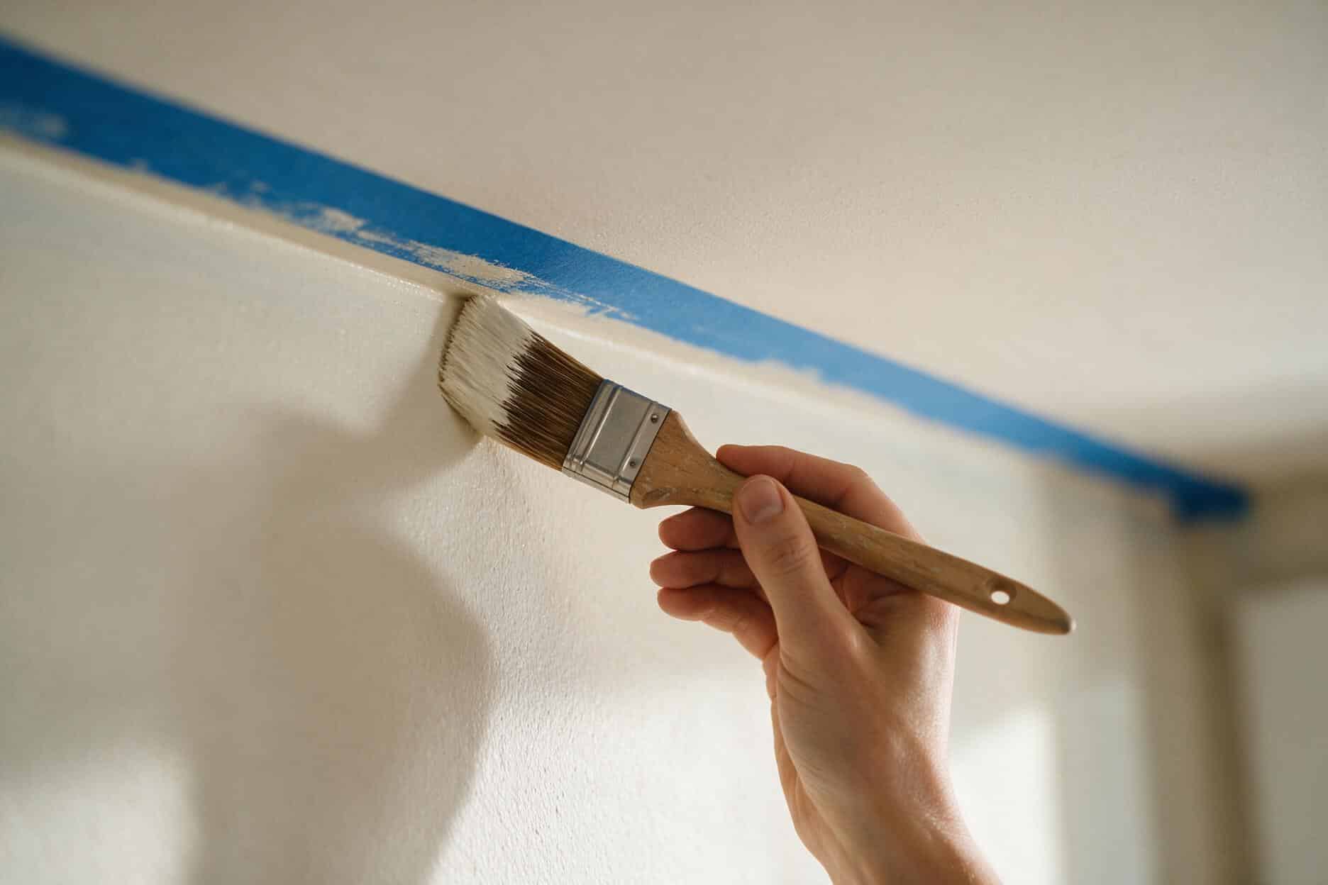 Hand holding an angled sash brush cutting in a crisp paint line along the wall-ceiling corner with painter's tape above