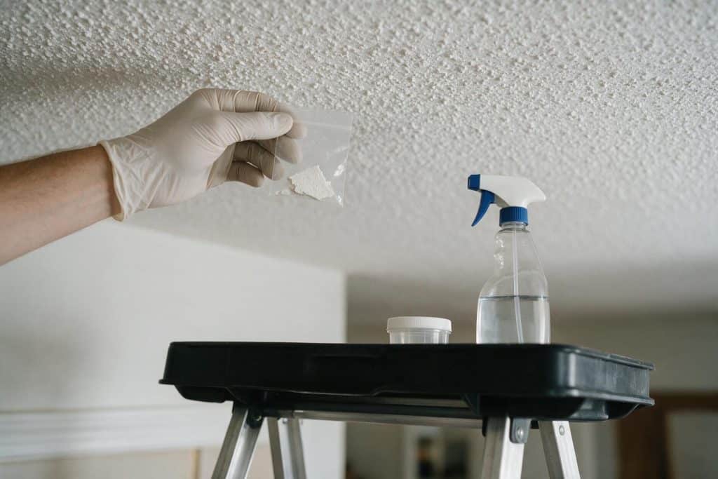 Gloved hand holding a sealed bag with a chip of popcorn ceiling material for asbestos testing