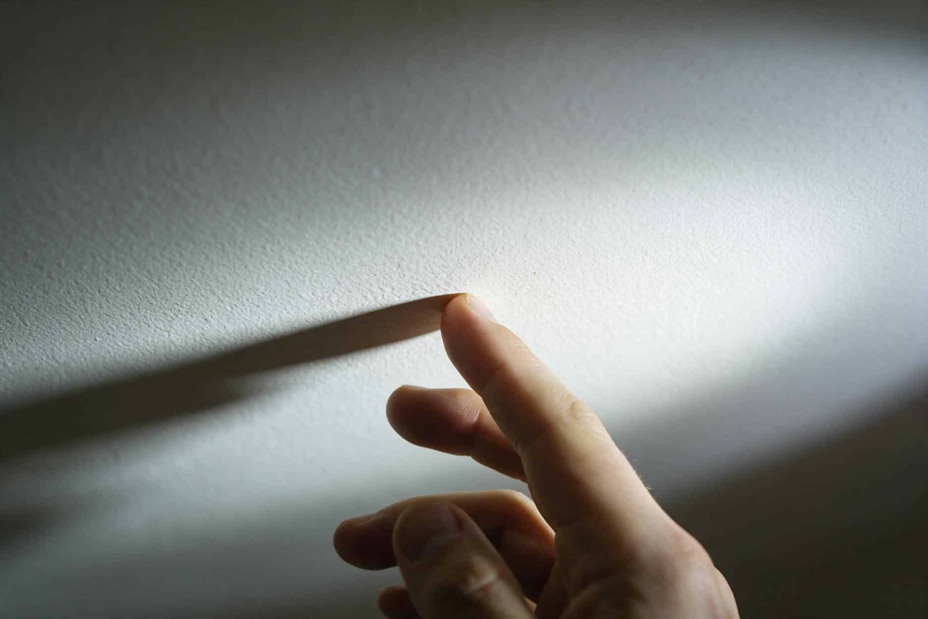 Close-up of fingertips pressing a ceiling to check for a soft spot, with raking light showing a faint texture change and no visible stain