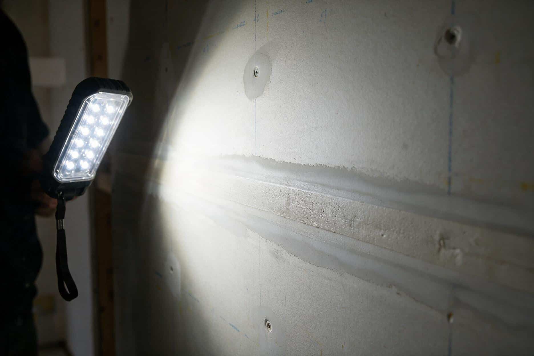 Work light held at a low raking angle across a sanded drywall wall, shadows revealing a faint ridge before primer