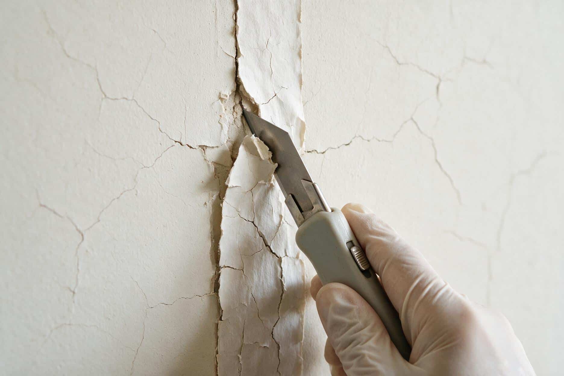 Gloved hand using a utility knife to score and lift old cracked tape off a drywall seam