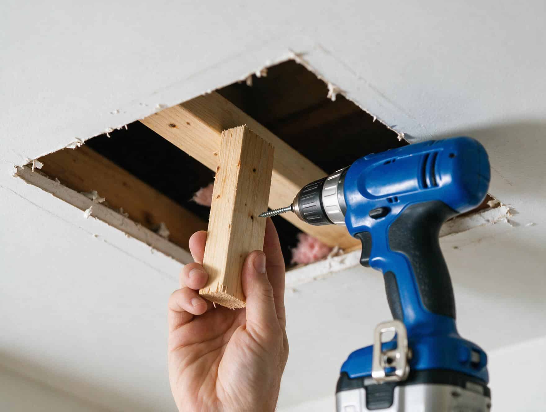 Hand holding a wood backing strip up into a ceiling cutout next to an exposed joist and insulation, drill ready to drive the screw