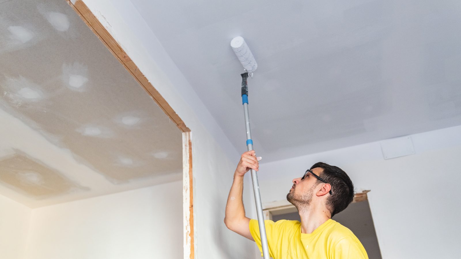Painter rolling white paint onto a ceiling with a roller on an extension pole, patched drywall visible in the next room