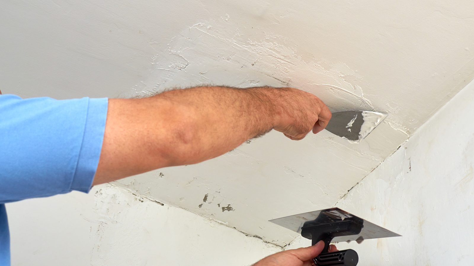 Hand spreading a thin coat of joint compound across a ceiling with a wide taping knife