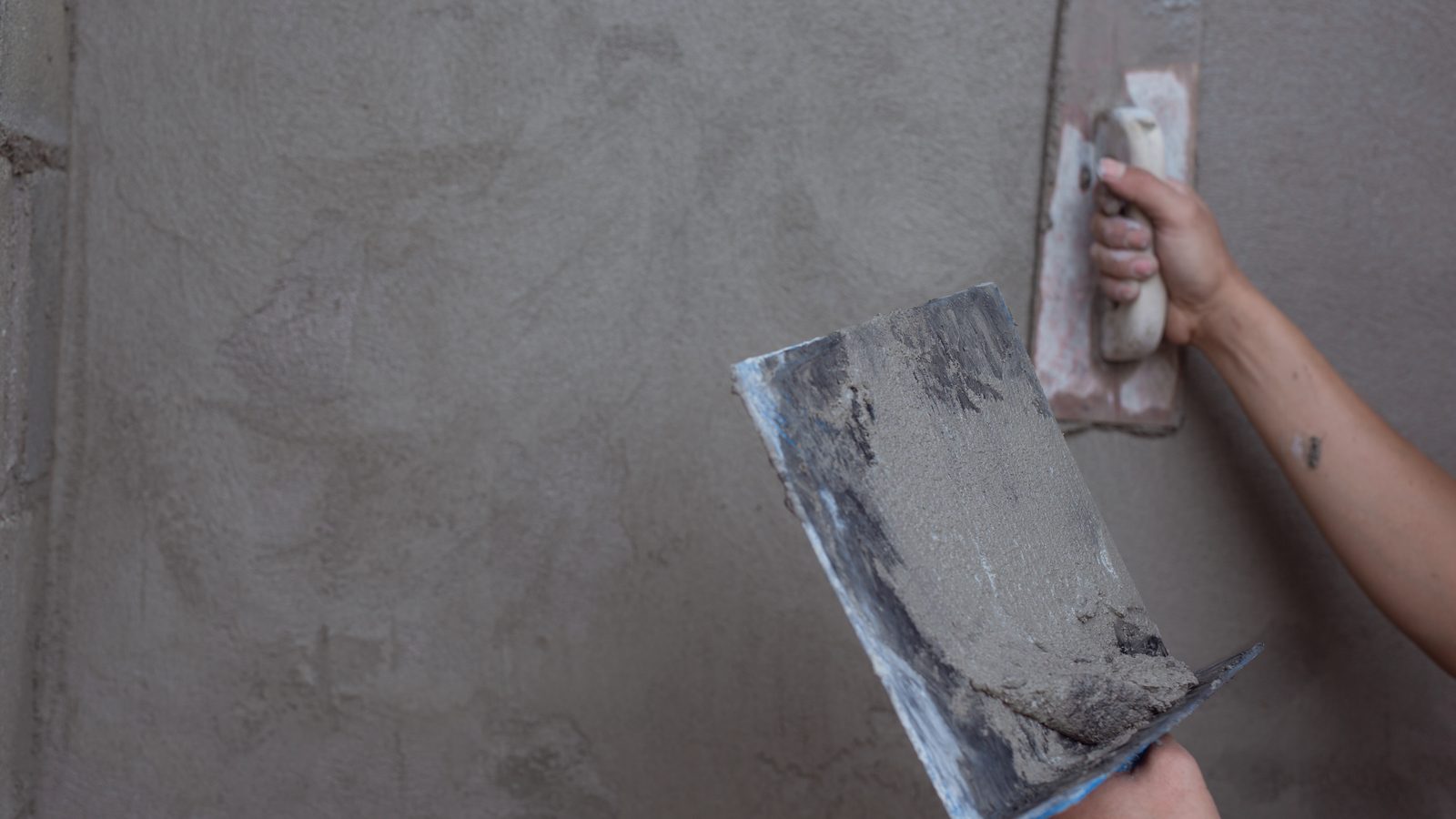 Finisher spreading a thin skim coat of joint compound over a wall with a wide knife to flatten texture before wallpaper