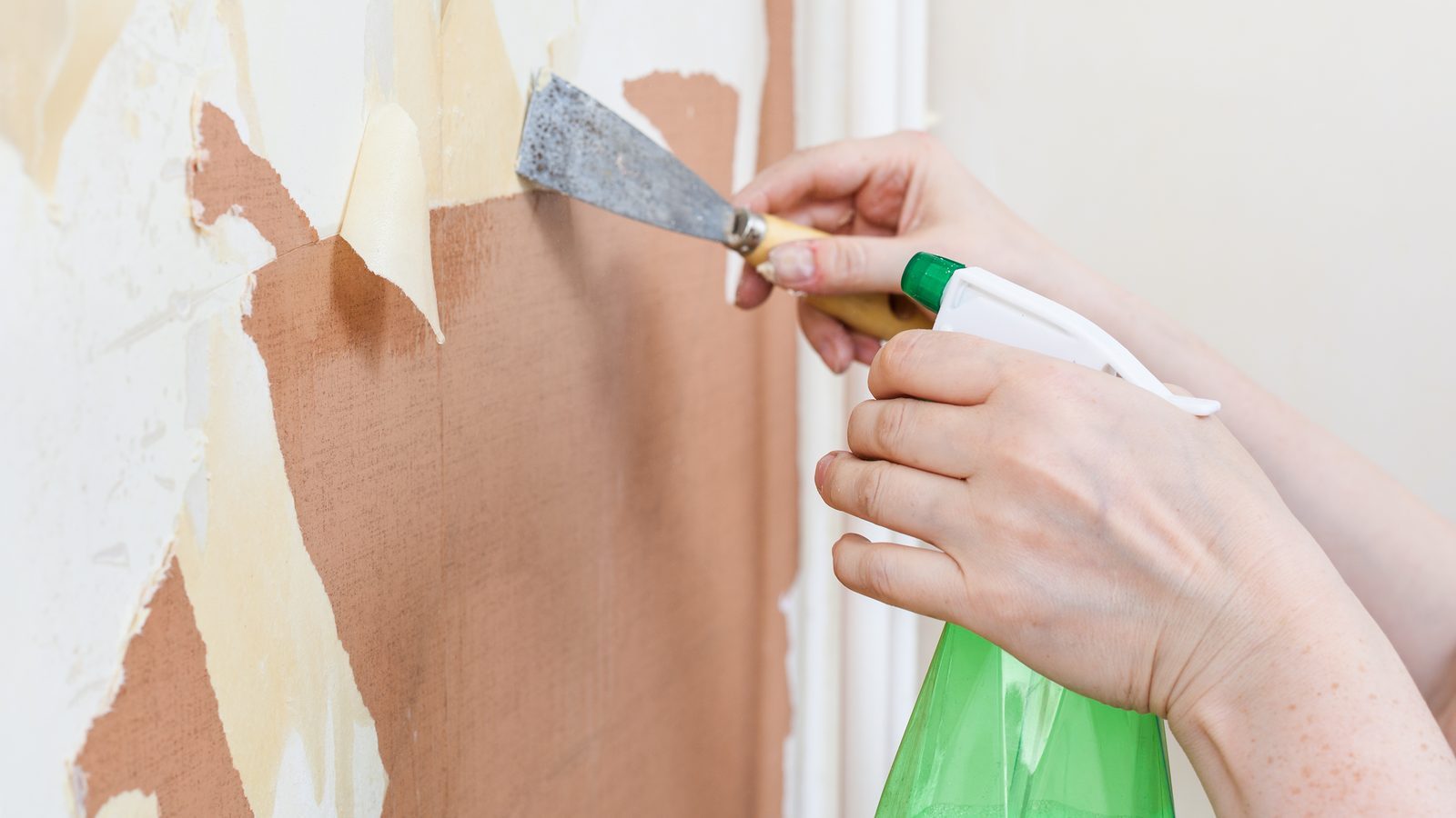 Hands holding a spray bottle and a putty knife scraping soaked wallpaper off a wall at a shallow angle
