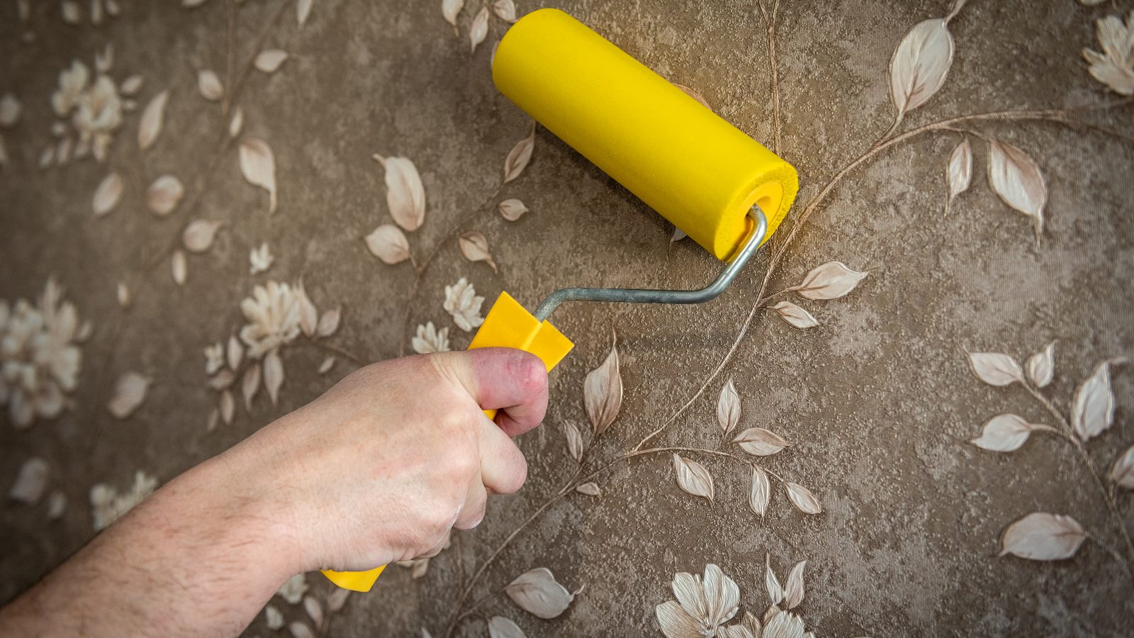 Hand pressing a foam roller across a freshly hung strip of patterned wallpaper to bond it flat to the wall