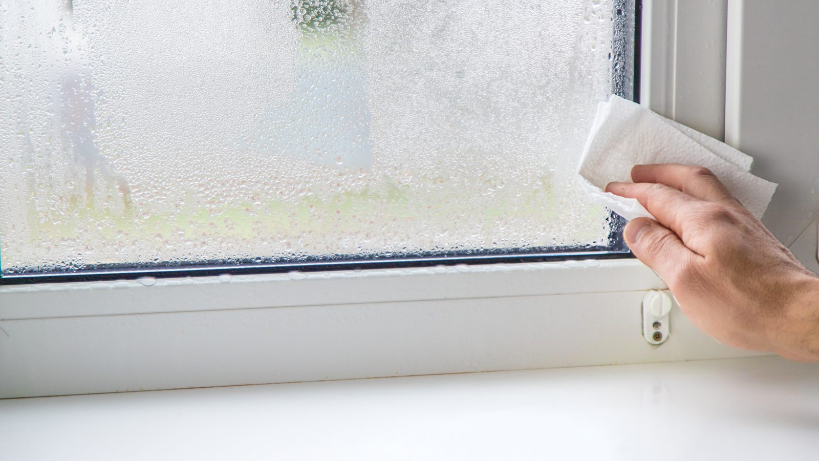Hand wiping heavy condensation off a white vinyl window pane with a paper towel, right at the bottom corner where the glass meets the sill.